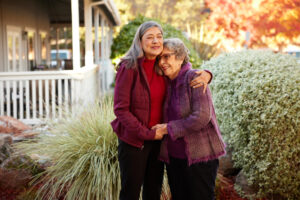 Two elderly women in purple jackets embrace and smile outdoors against autumn foliage and ornamental grass.