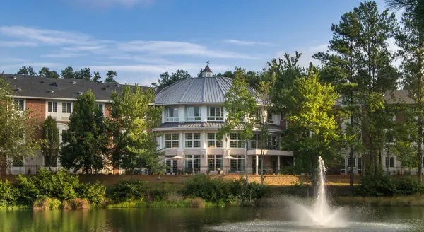 A serene lakeside setting with a charming building, inviting greenery, and a gentle fountain creates a welcoming atmosphere.