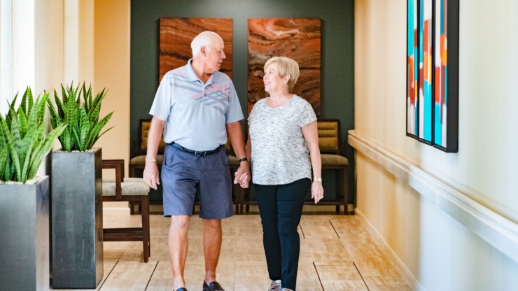 A couple strolls hand-in-hand down a welcoming corridor, surrounded by vibrant artwork and greenery.