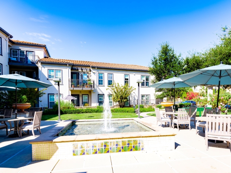 A sunny courtyard featuring cozy seating, a tranquil fountain, and vibrant gardens, inviting community connections.