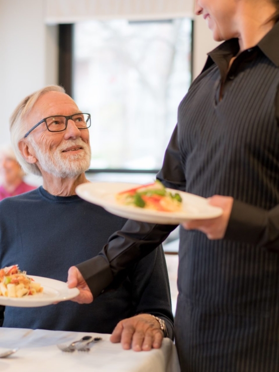A cheerful dining scene where a resident enjoys a meal served with care and attention in a warm community atmosphere.