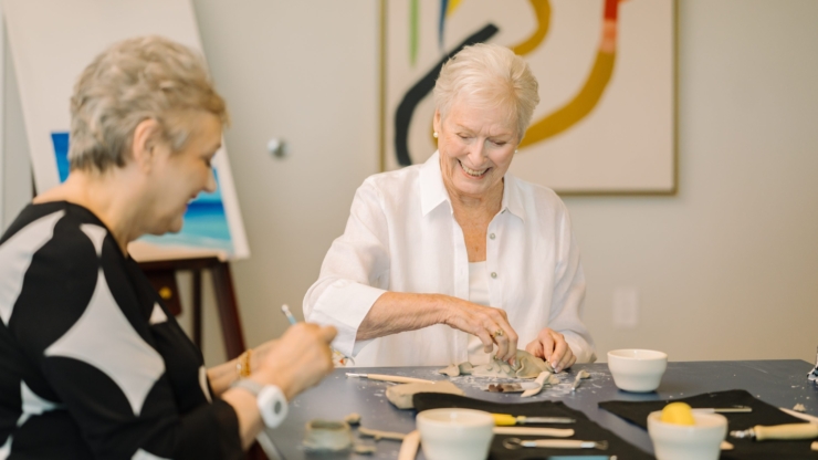 Two smiling women engage in a creative activity, surrounded by art and a warm, inviting atmosphere.