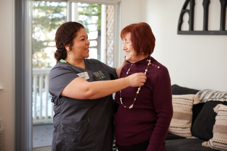 A caregiver warmly assists a resident with a necklace, radiating joy and community spirit in a cozy setting.