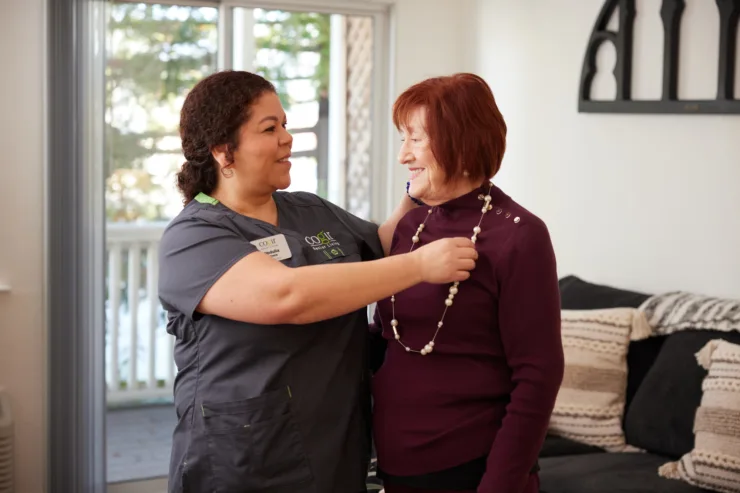 A caregiver warmly assists a resident with a necklace, radiating joy and community spirit in a cozy setting.