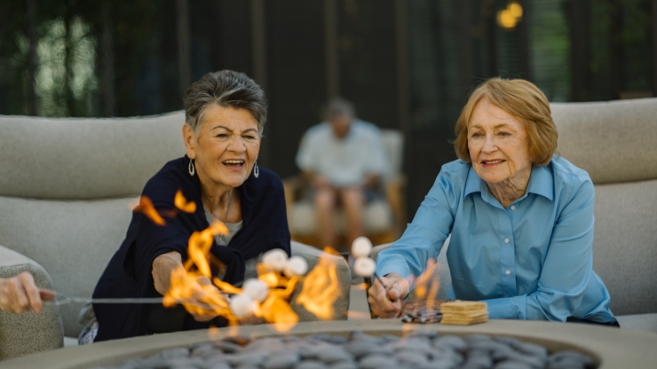 Two joyful women enjoy roasting marshmallows by a cozy fire, surrounded by a welcoming atmosphere.