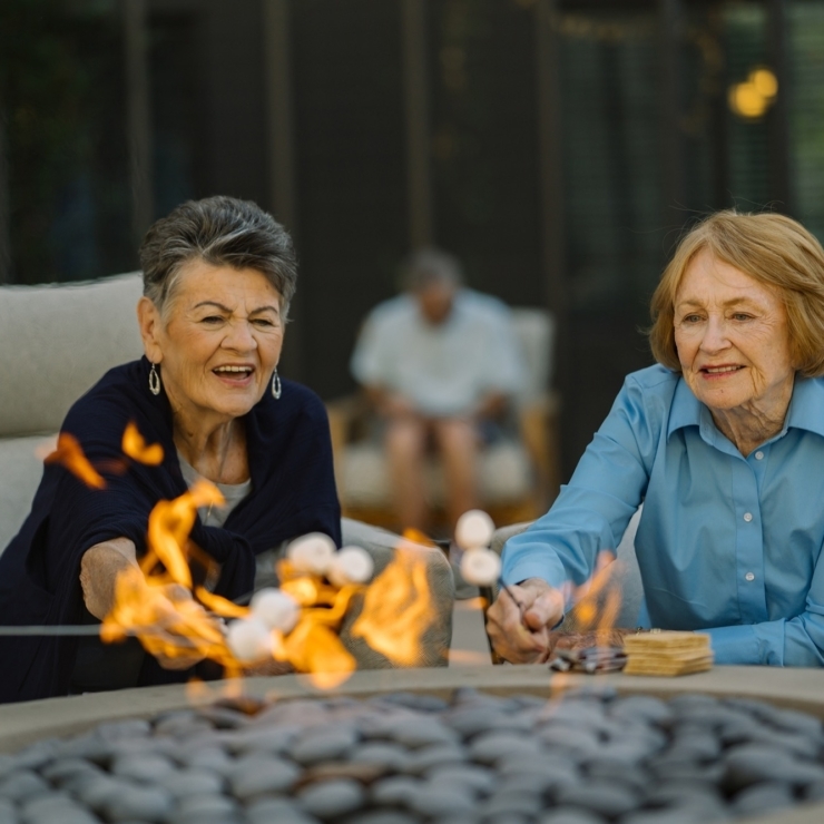 Two joyful women enjoy roasting marshmallows by a cozy fire, surrounded by a welcoming atmosphere.