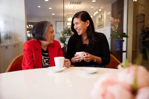 Two women share a joyful moment over coffee, surrounded by fresh blooms and a welcoming atmosphere.