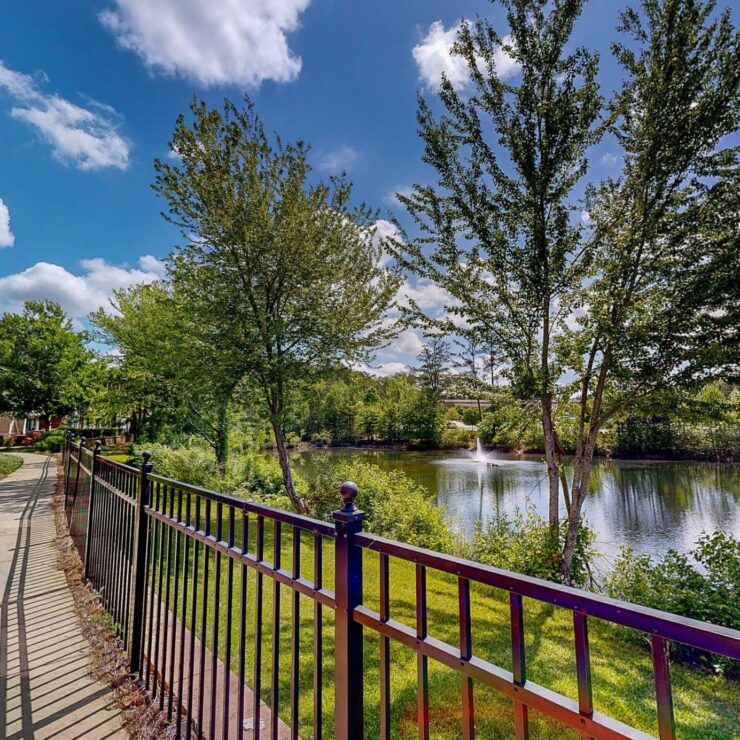 A serene pathway winds alongside a tranquil pond, framed by lush greenery and bright blue skies.
