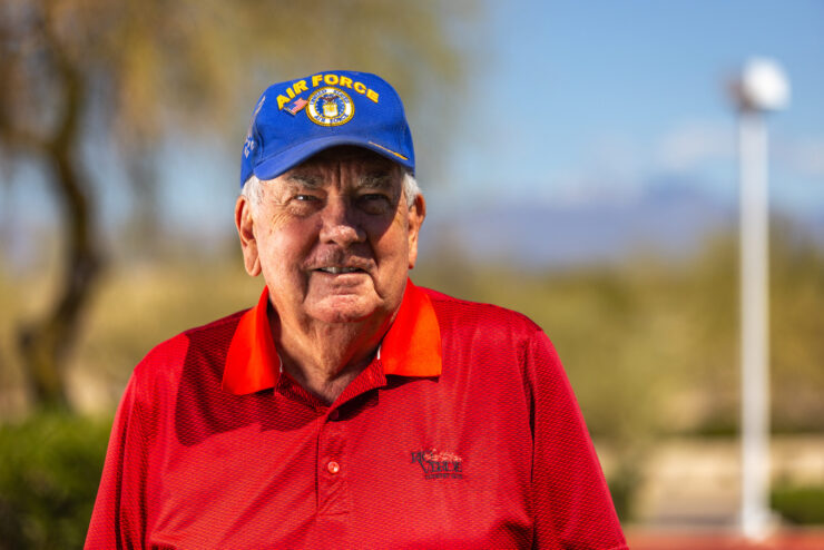 A smiling gentleman in a vibrant red shirt and Air Force cap stands warmly, embodying community spirit.