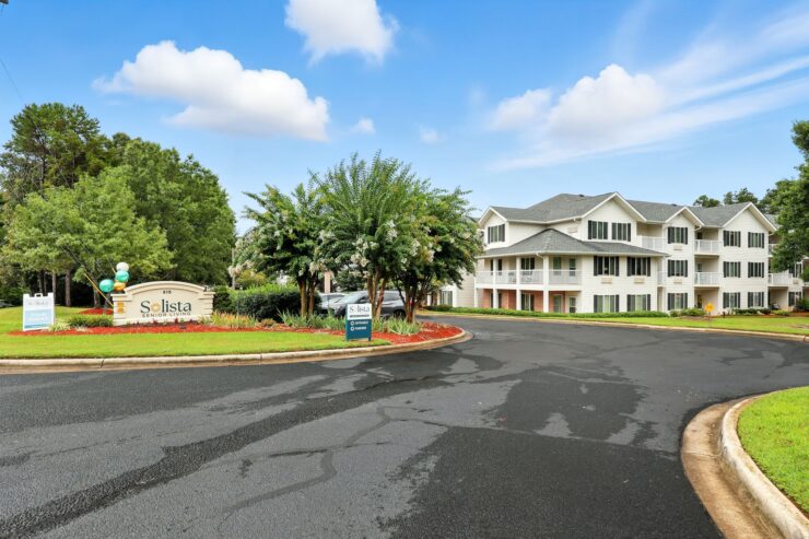 A welcoming entrance to Solista Senior Living, surrounded by lush greenery and a bright sky, evokes community spirit.