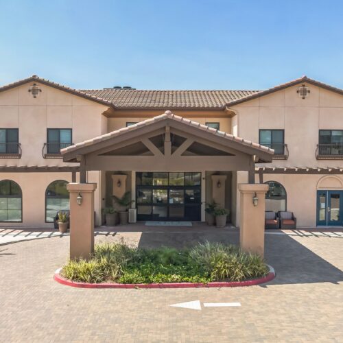 Well-maintained retirement home entrance with shaded seating and landscaped greenery under clear skies.