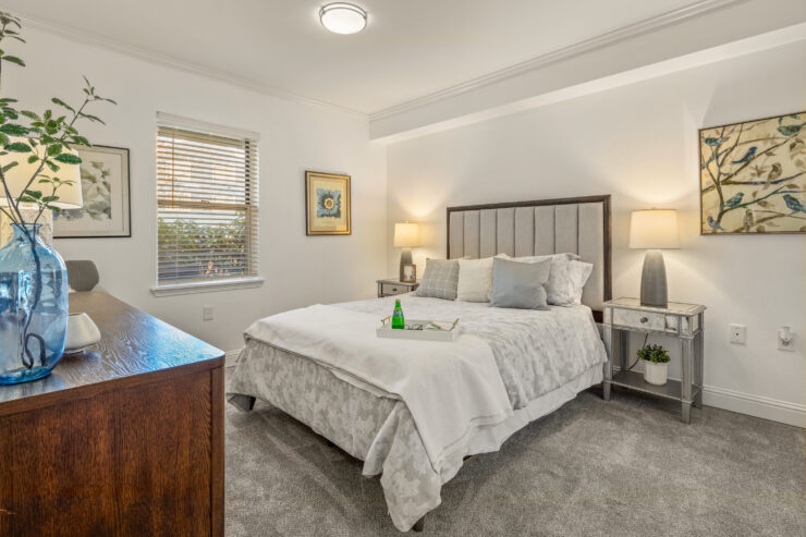 Sunlit bedroom with plush bedding, soft carpet, and calming artwork for relaxing comfort.