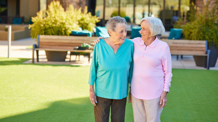 Two elderly women sharing a warm moment in a sunny, inviting outdoor courtyard space.