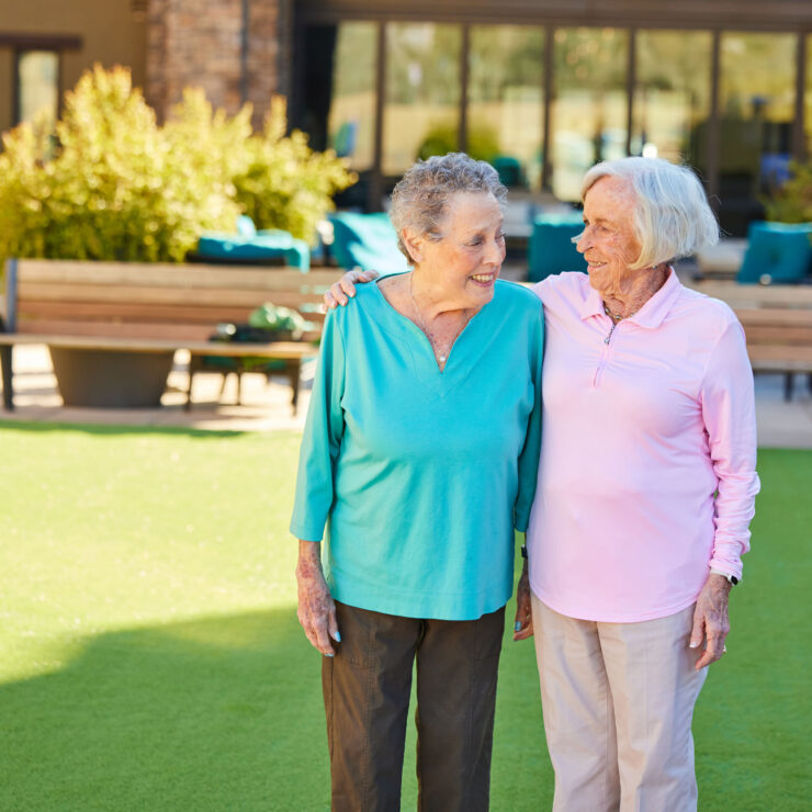 Two elderly women sharing a warm moment in a sunny, inviting outdoor courtyard space.