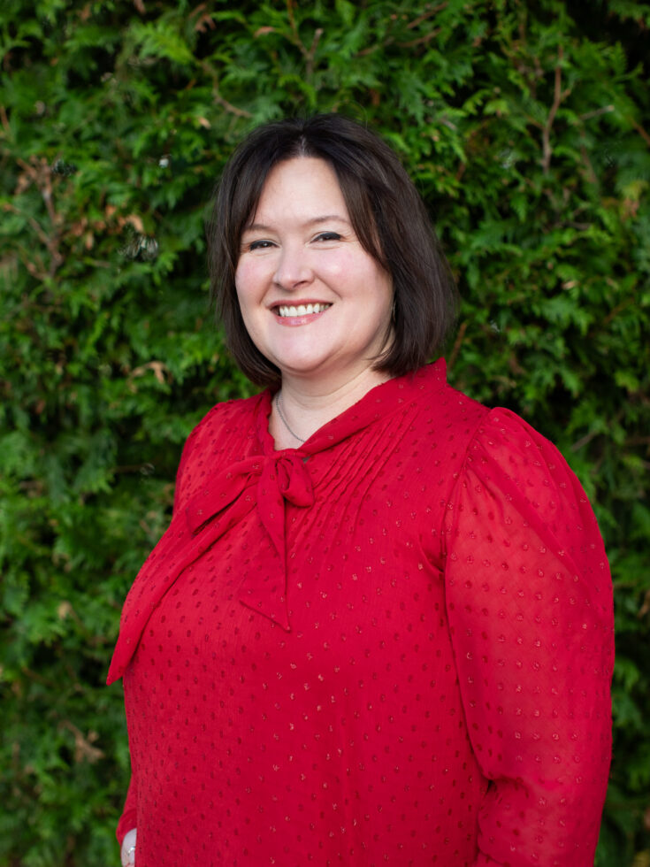 A warm smile radiates from a friendly woman against a lush green backdrop, embodying community spirit.
