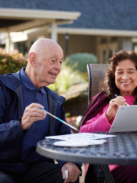 Two seniors enjoy painting together outdoors, sharing smiles in a peaceful garden setting.