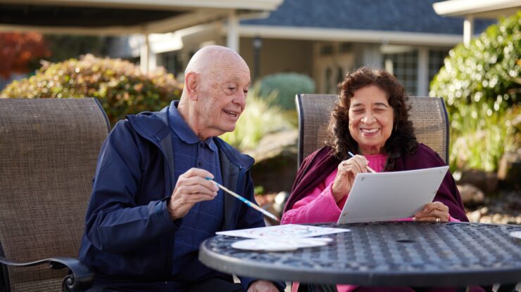Two seniors enjoy painting together outdoors, sharing smiles in a peaceful garden setting.