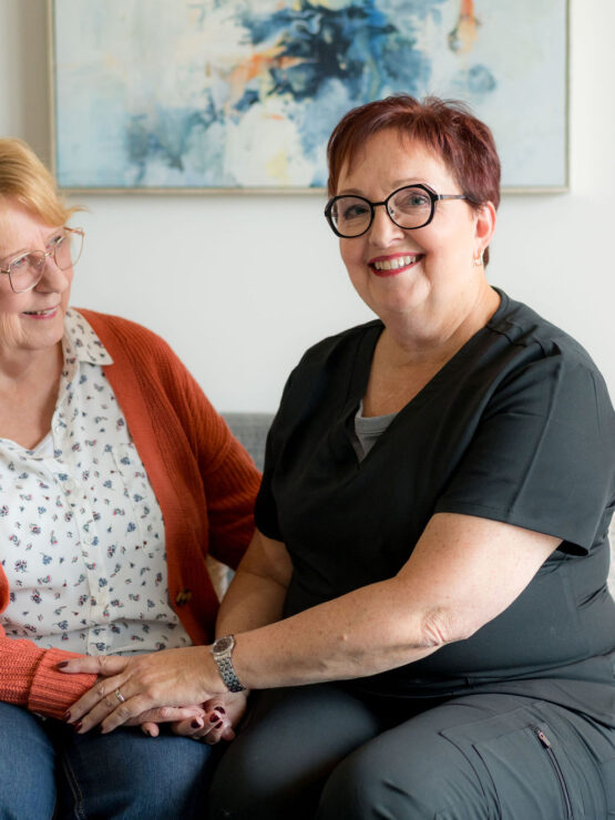 Two smiling women share a warm moment on a sofa, reflecting care and companionship.