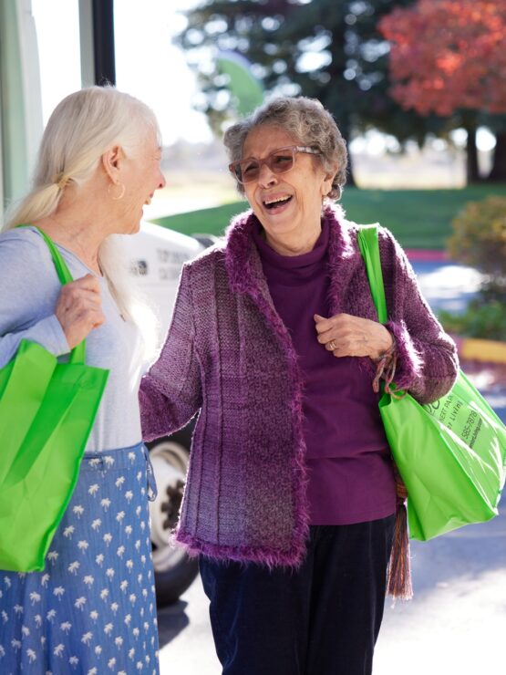 Two elderly women smiling and chatting outdoors, carrying bright green bags near a bus.