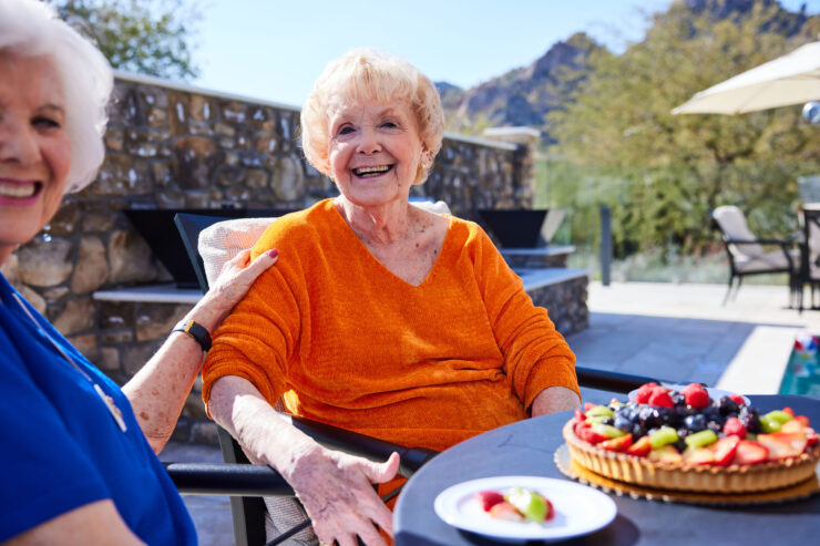 Two smiling seniors enjoy fresh fruit tart together in a bright, welcoming outdoor space.