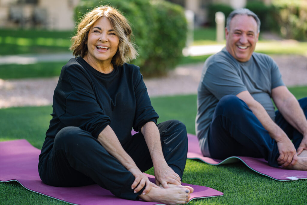 Two smiling individuals practice yoga on mats in a vibrant, green outdoor setting, radiating joy and community.
