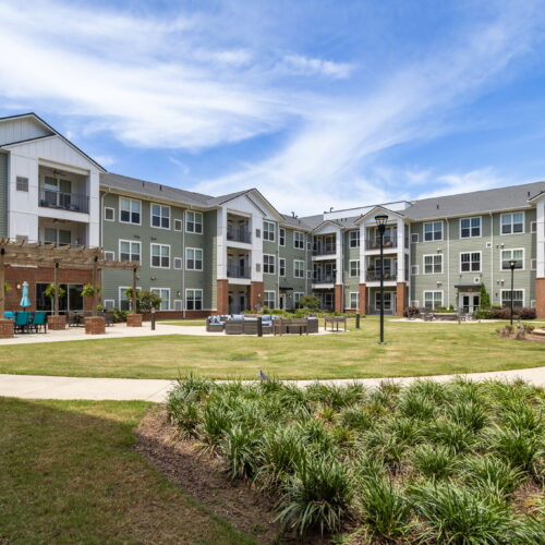 Spacious retirement community courtyard with seating areas, greenery, and a bright blue sky.