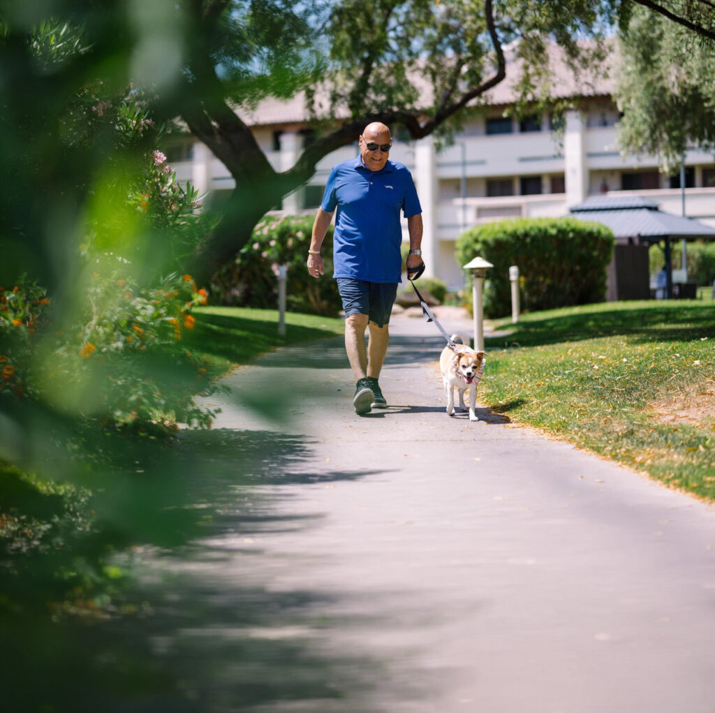A man strolls along a sunlit path with his dog, surrounded by vibrant greenery and friendly community vibes.