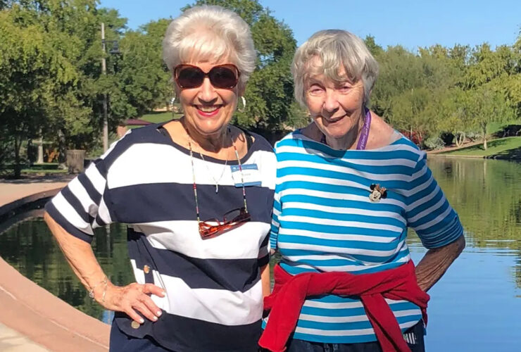 Two cheerful women in striped shirts enjoy a sunny day by a serene pond, embracing community spirit.