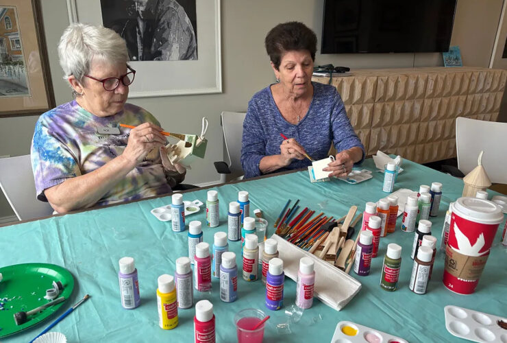 Two women painting small wooden crafts at a vibrant, welcoming community table.