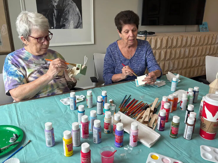 Two women painting small wooden crafts at a vibrant, welcoming community table.