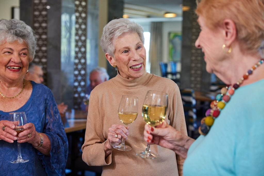 Three senior women enjoying conversation and wine in a welcoming community lounge.