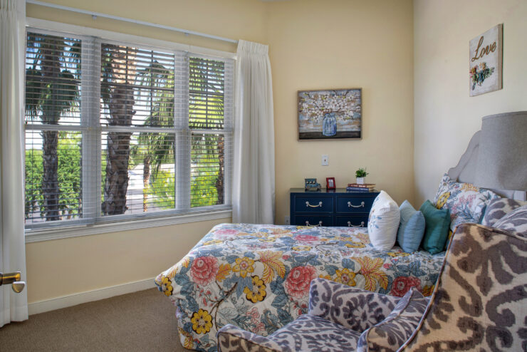 Sunlit bedroom with floral bedding, comfortable armchair, and a view of palm trees outside.