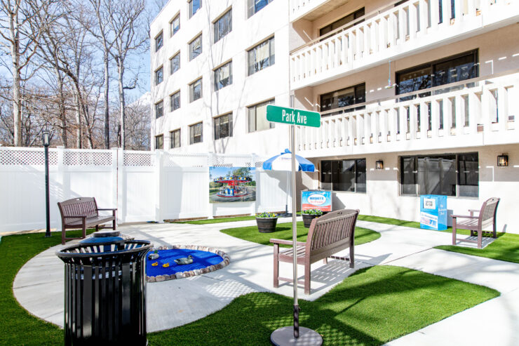 Sunlit outdoor seating area with benches, greenery, and community signs at a retirement home.