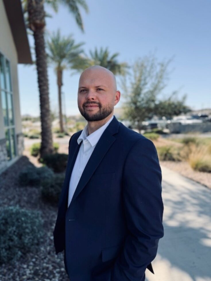 Friendly staff member standing outdoors near landscaped pathways and palm trees.
