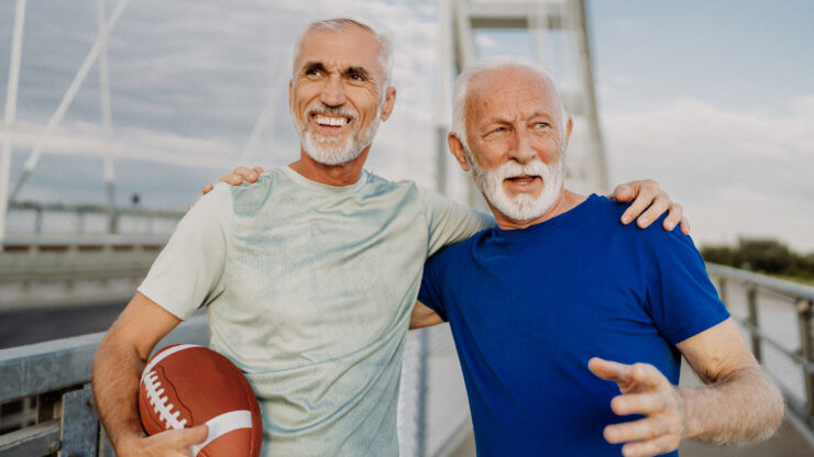 Two senior men smiling and enjoying an active, friendly moment outdoors on a bridge.