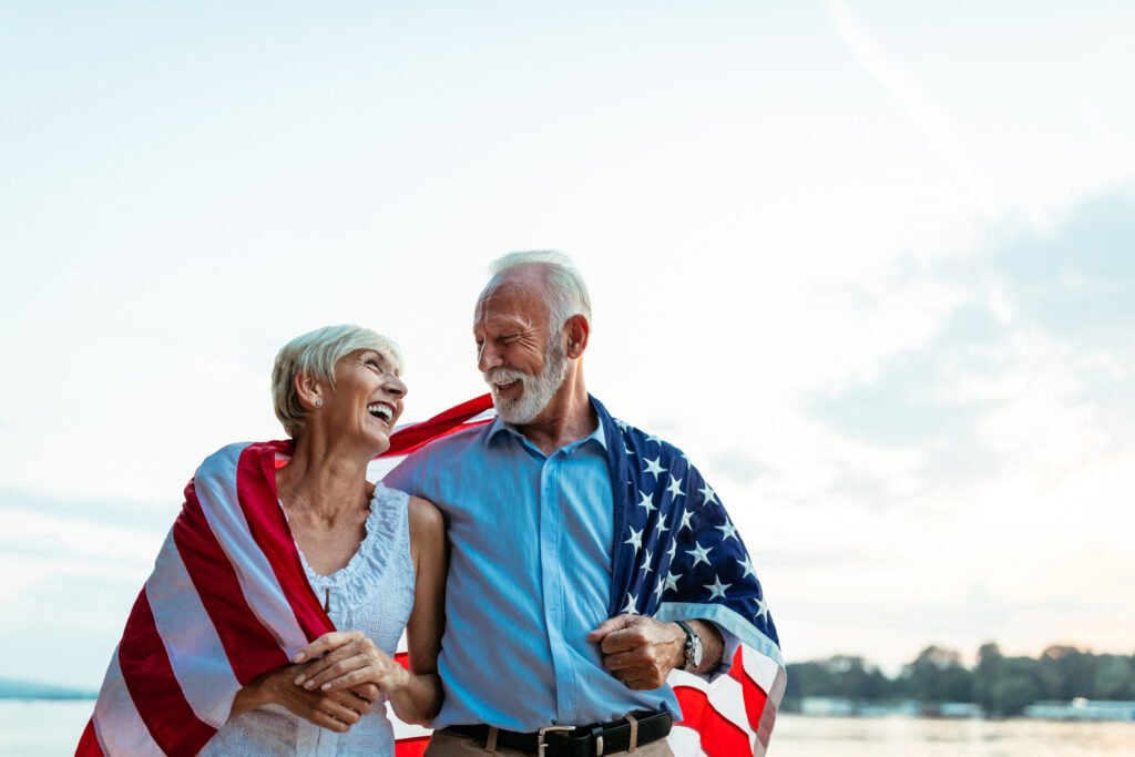 Elderly couple wrapped in an American flag, smiling together by a peaceful waterfront.