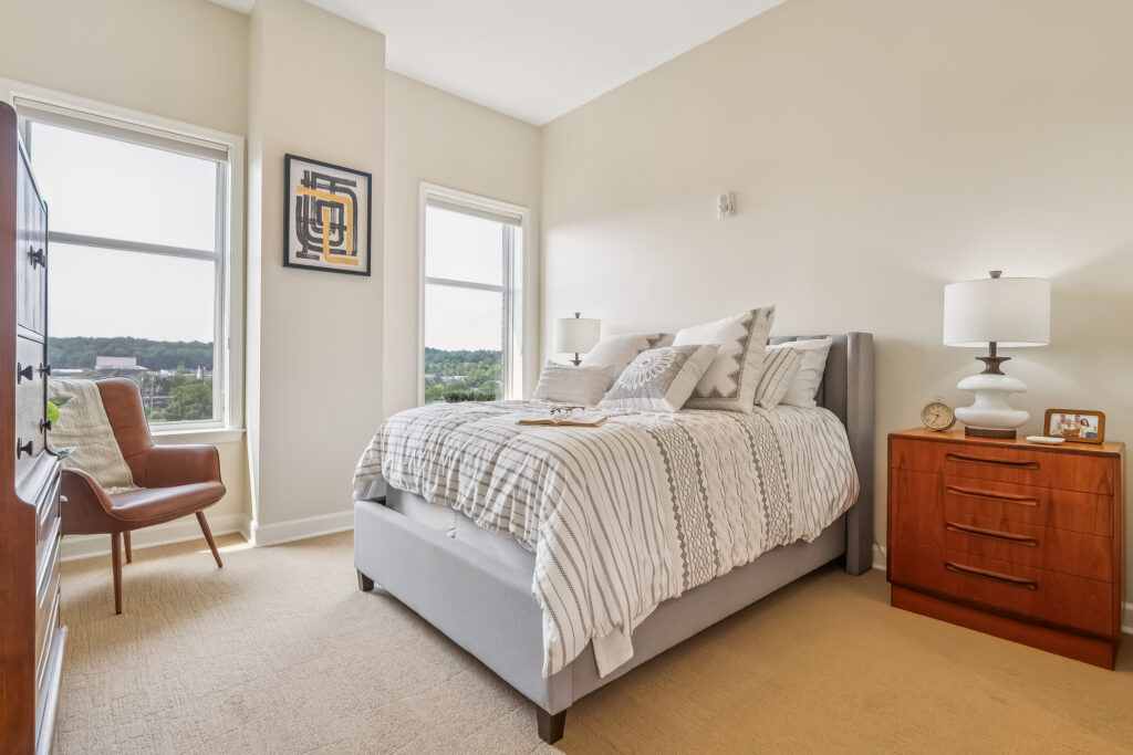 Sunlit bedroom with warm wood furniture, soft bedding, and large windows overlooking greenery.