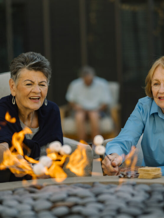Two senior women enjoy roasting marshmallows together around a fire pit in a relaxed outdoor setting.