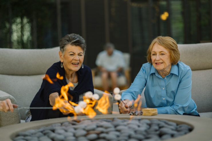 Two senior women enjoy roasting marshmallows together around a fire pit in a relaxed outdoor setting.