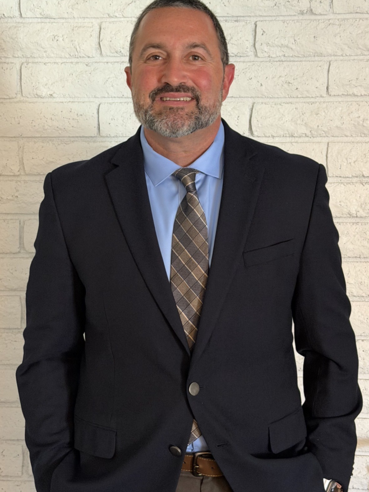 Smiling professional man in suit stands relaxed against a light brick wall, welcoming atmosphere.
