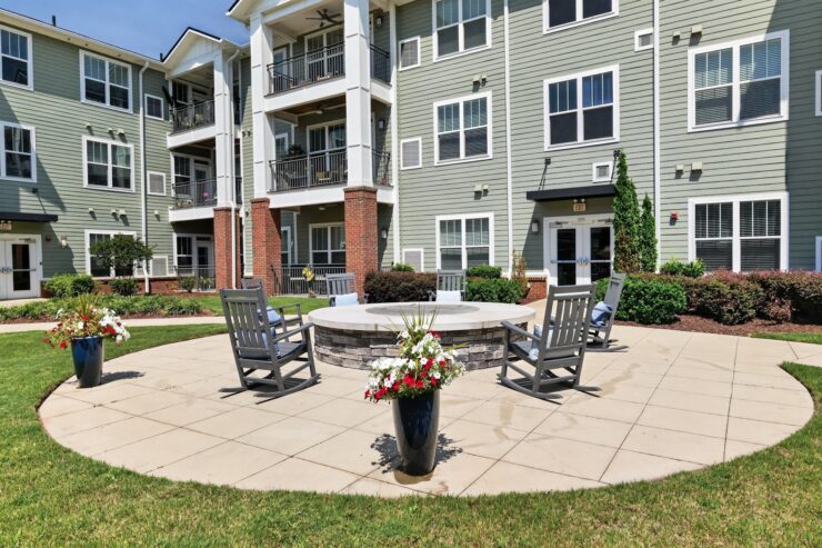 Outdoor seating area with a firepit, surrounded by greenery and a welcoming community building.