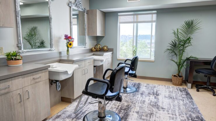 Bright salon area with styling chairs, large mirrors, plants, and natural light for residents' comfort.