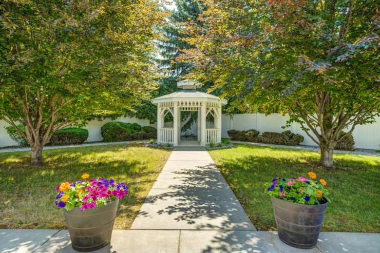 Tranquil garden with vibrant flowers, shaded trees, and a charming white gazebo pathway.