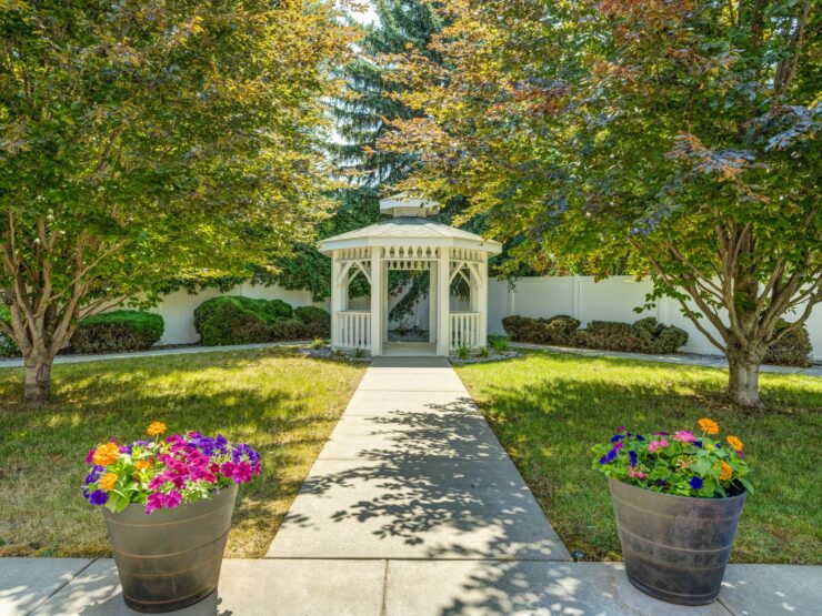 Tranquil garden with vibrant flowers, shaded trees, and a charming white gazebo pathway.