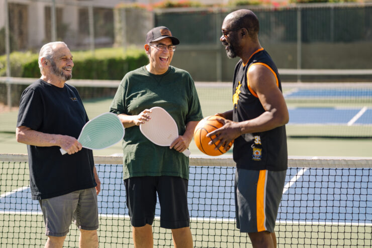 Three seniors laugh and chat while holding sports equipment on a sunny court, enjoying friendship.