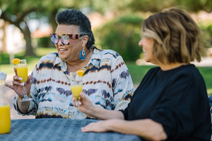Two women enjoy a sunny outdoor gathering, sharing drinks and laughter in a peaceful garden.