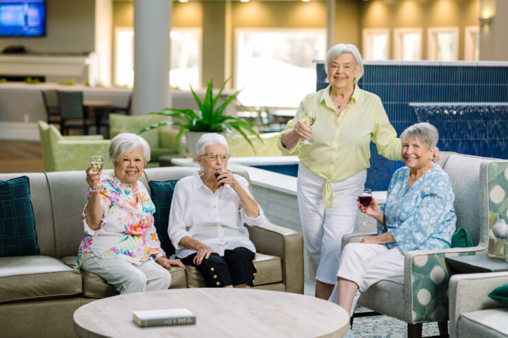 Four senior women enjoying drinks together in a bright, inviting common area with comfortable seating.
