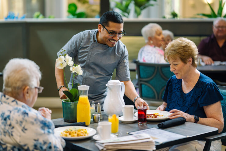 Caregiver serves breakfast to smiling seniors enjoying a bright dining area with plants.