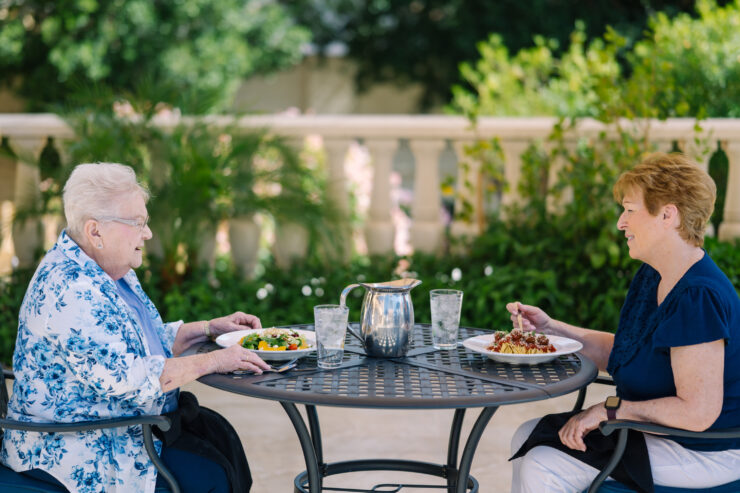 Two women enjoy a meal together at a garden patio surrounded by lush greenery.