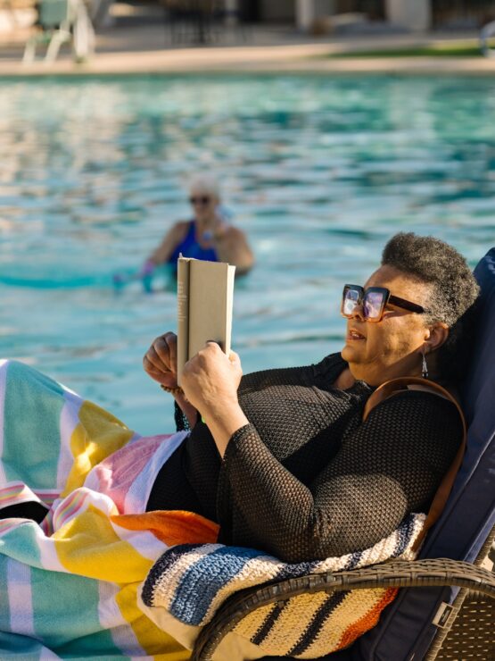 Resident enjoys reading by the pool while another exercises in the water, embracing wellness.